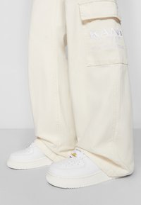Pantalones cargo color crema con bolsillos laterales y logo bordado, combinados con zapatillas blancas con detalles perforados y una suela texturizada.