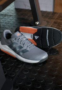Zapatillas deportivas grises con parte superior de malla, suela de goma negra y naranja, tres franjas negras en el lateral, tejido texturizado y diseño con cordones.