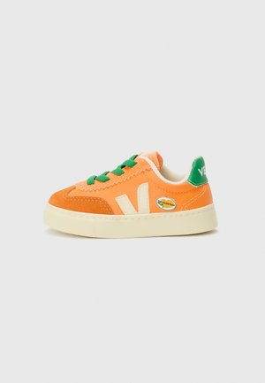 Pomarańczowo-beżowy sneaker z zielonymi sznurowadłami i zawieszką na pięcie, z białym logo „V” i małą plakietką „wegańską” po boku.