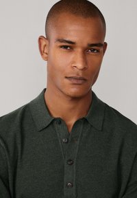 Tamnozeleno pleteno polo majice s teksturiranim završetkom, koja ima klasičan ovratnik i tri dugmeta na prednjem dijelu.