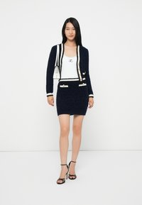 Donna in piedi contro uno sfondo bianco, indossa una gonna blu navy con texture e un cardigan con rifiniture bianche, una maglietta bianca, sandali neri con tacco alto e una borsa a tracolla bianca.