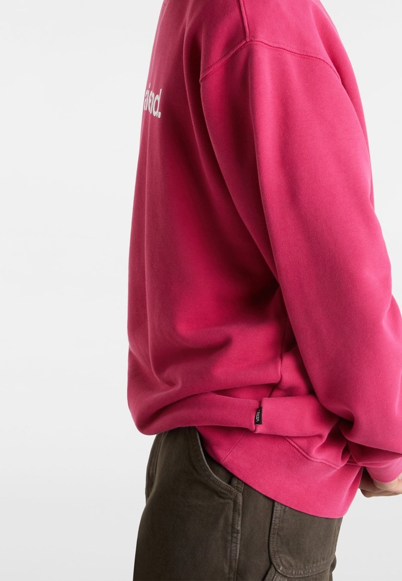 Personne portant un sweat-shirt rose vif avec une petite étiquette Vans sur l'ourlet latéral, associée à un pantalon marron foncé, vu de profil.