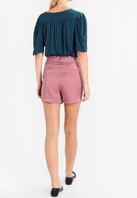Blusa teal con maniche a sbuffo, abbinata a pantaloni corti rosa a vita alta. Scarpe nere senza lacci. Design semplice, texture liscia.