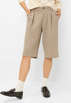 Kvinde iført beige knælang skræddersyet shorts, beige striktop, hvide sokker og mørkebrune loafers står med hænderne i lommerne.