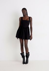 Melnā mini kleita ar kvadrātveida kakla izgriezumu, pieguļošu augšdaļu un platām svārku daļām. Modelis valkā garas, melnas, brīvas botas ar spīdīgu tekstūru.