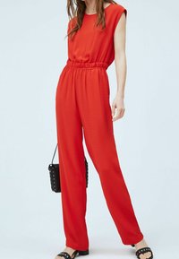 Combinaison rouge avec manches courtes, taille élastique et coupe ample. Portée avec un sac à main noir clouté et des sandales. Texture lisse.