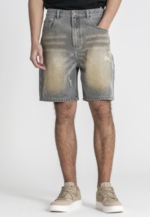 Mann trägt ausgewaschene graue Jeansshorts mit beigen Flicken, schwarzes Shirt und beige Sneakers, steht mit einer Hand in der Tasche.