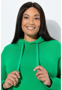 Zelená mikina s kapucí, přední kapsou, nastavitelnými stahovacími šňůrkami a měkkou texturou. Model drží stahovací šňůrky s kovovými koncovkami.