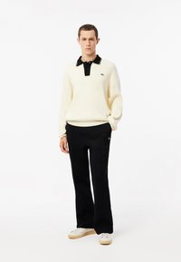 Suéter de canalé color crema con un cuello negro y una tapeta de tres botones, combinado con pantalones negros y zapatillas blancas, que presentan un logo verde.