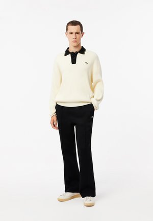 Suéter de canalé color crema con un cuello negro y una tapeta de tres botones, combinado con pantalones negros y zapatillas blancas, que presentan un logo verde.