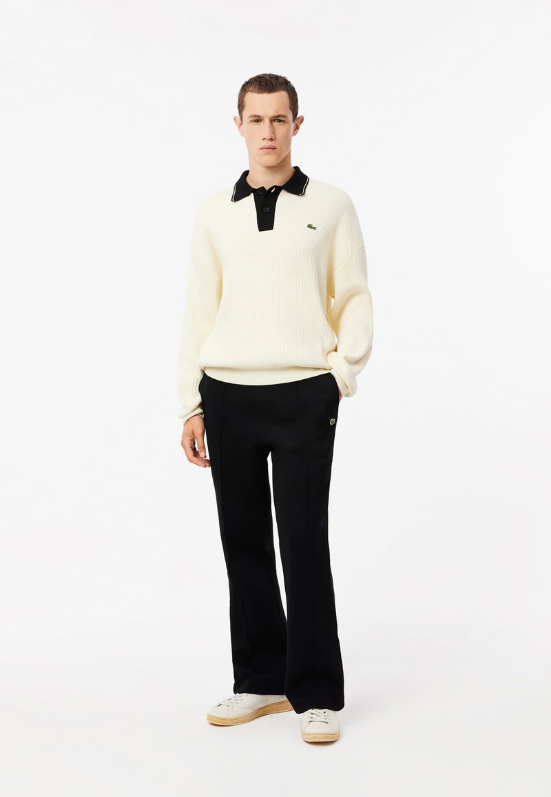 Suéter de canalé color crema con un cuello negro y una tapeta de tres botones, combinado con pantalones negros y zapatillas blancas, que presentan un logo verde.