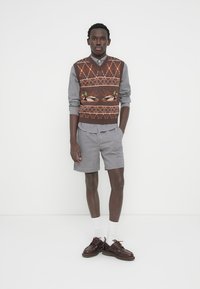 Άνδρας που στέκεται, φορώντας καφέ γιλέκο με μοτίβο πάπιας πάνω από καρό πουκάμισο, συνδυασμένο με σορτς και καφέ δερμάτινα παπούτσια.