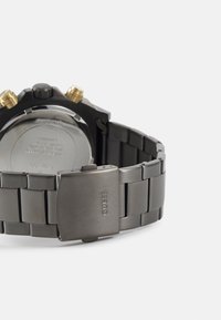 Reloj de metal negro con un acabado brillante, acentos en oro en la corona y un cierre de acero inoxidable. Presenta una pulsera de estilo eslabonado con textura.