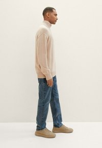 Jersey de punto beige con cuello alto, combinado con jeans azules y zapatillas de color crema claro. El jersey cuenta con puños y dobladillo acanalados, y tiene un corte relajado.