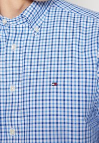 Hellblau-weiß kariertes Button-Down-Hemd mit Kragen, das ein kleines Logo-Detail in Rot, Weiß und Blau aufweist.
