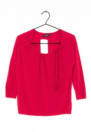 Jersey rojo de manga larga con escote redondo y lazos laterales, colgado en una percha negra sobre un fondo blanco.