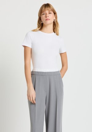 Donna bionda che indossa una maglietta bianca aderente a maniche corte e pantaloni grigi a gamba larga a vita alta, in piedi con una mano in tasca.