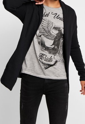 Cardigan nero con apertura frontale, tessuto testurizzato. Indossato sopra una t-shirt grigia con grafica che presenta un design di teschio nero e ali. Jeans slim-fit.