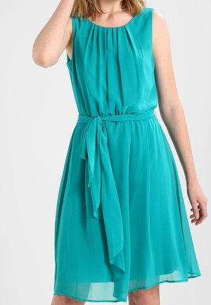 Kobieta ubrana w turkusową sukienkę bez rękawów z plisowanym gorsetem i przewiązanym paskiem w talii, stojąca na jasnym, jednolitym tle.
