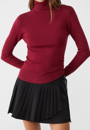 Femme portant un pull col roulé bordeaux ajusté et une jupe plissée noire taille haute, debout les mains dans le dos.