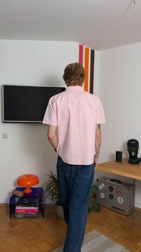 Camicia rosa a maniche corte, jeans blu, in piedi in una stanza con una TV a schermo piatto e strisce colorate sulle pareti. Presenta una lampada arancione e piante.