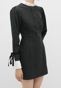 Robe noire à carreaux avec manches longues bouffantes, grands boutons circulaires sur le devant, poignets plissés blancs et col blanc.