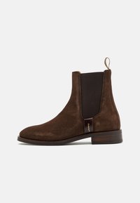 Bruine suède enkellaars met elastische zijpanelen en een treklabel. Heeft een houten hak en een gouden logo-accent aan de zijkant.