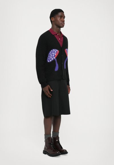 Cardigan negru cu desene de ciuperci violet și roșu peste un polo bordeaux, pantaloni scurți negri până la genunchi, șosete gri și botine maro cu șireturi.