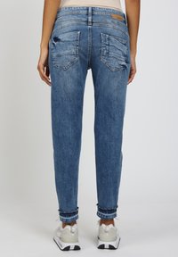 Person in blauen hochgekrempelten Jeans und weißen Sneakers, entspannt mit den Armen an den Seiten auf einem einfarbigen Hintergrund stehend.