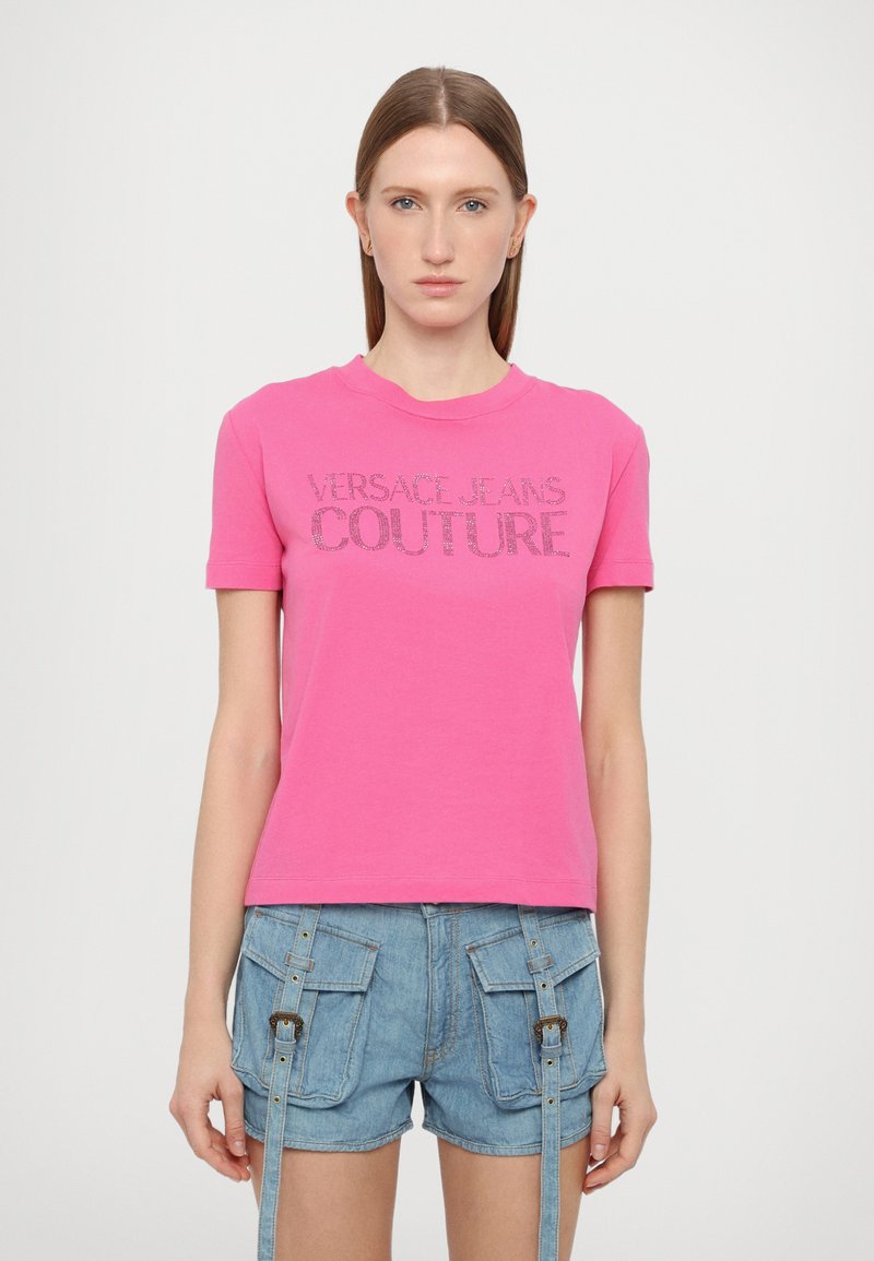 Rožinė medvilninė marškinė su „VERSACE JEANS COUTURE“ tekstu krištolais, apvaliu iškirpimu, trumpomis rankovėmis, derinama su džinsiniais šortais.