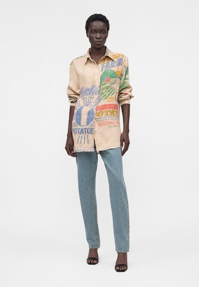 Γυναίκα που φοράει φαρδιά μπεζ μπλούζα με πολύχρωμο σχέδιο αγροκτήματος και πατάτας, ανοιχτόμπλε τζιν και μαύρες ανοιχτές γόβες, στέκεται μπροστά σε λευκό φόντο.