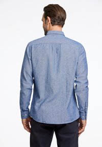 Camicia in chambray blu con maniche lunghe, polsini a bottone e dettaglio di piega sulla schiena. Texture liscia, motivo di tessitura delicato, orlo curvo.