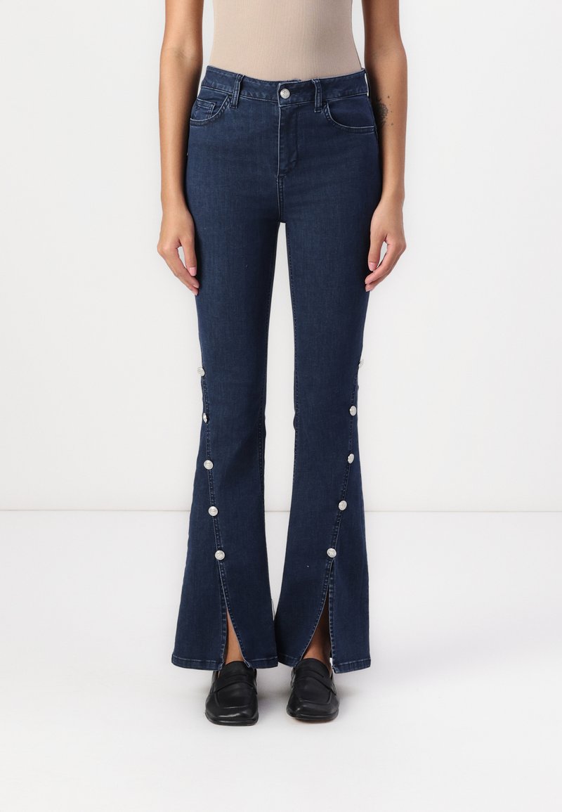 Jeans de campana de color azul oscuro con cintura alta, bolsillos delanteros, detalles de botones a lo largo de las costuras laterales y dobladillos divididos en los tobillos.