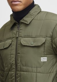 Olijfgroene gewatteerde jas met een rits aan de voorkant, drukknopen op de borstzakken, gemaakt van nylon en een label met de tekst "11 PROJECT" op de rechteronderkant.