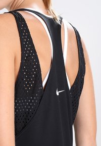 Canottiera sportiva nera con schiena in rete e ritagli circolari, sovrapposta a un reggiseno sportivo bianco. Logo Nike visibile sul retro.