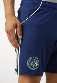 Tumes sinised sportlikud lühikesed püksid helesiniste külgriidega ja ümmarguse tikandiga logoga, millel on klassikaline figuur. Sile, veniv kangas.