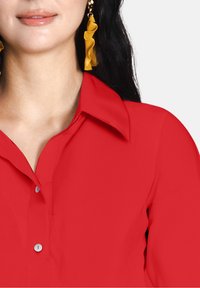 Camicia rossa con un colletto classico, frontale con bottoni e una texture morbida. Orecchini arricciati dorati visibili, che migliorano l'aspetto generale.