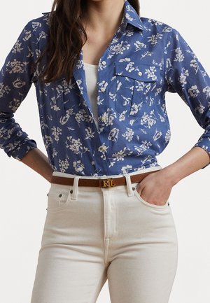 Femme portant une chemise bleue à fleurs boutonnée par-dessus un top blanc, rentrée dans un pantalon blanc taille haute avec une ceinture marron avec des initiales dorées.