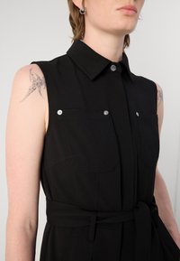 Vestido negro sin mangas con collar, botones frontales, dos bolsillos en el pecho y un cinturón de tela en la cintura. Textura suave, diseño minimalista.