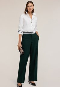 Camicia bianca con maniche a righe nere, abbinata a pantaloni larghi verdi su misura. Gli accessori includono una pochette argentata e tacchi.