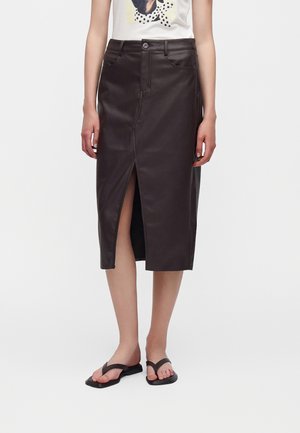 Femme portant une jupe midi en cuir noir avec une fente devant, un t-shirt blanc à motif graphique, et des sandales plates noires sur un fond blanc uni.