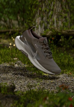 Hnědé a bílé běžecké boty Nike na trail s technologií Gore-Tex, vznášející se ve vzduchu nad štěrkovou cestou v lesním prostředí.