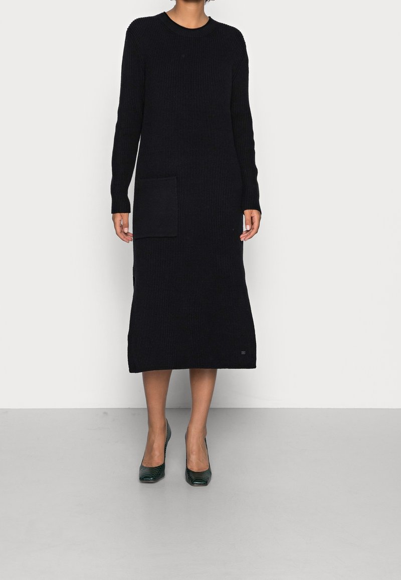 Robe longue noire en maille avec texture côtelée, encolure ronde et poche latérale. Portée avec des talons verts, mettant en valeur une silhouette ajustée.