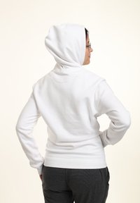 Sudadera blanca de algodón con capucha redondeada, puños y dobladillo acanalados; el modelo lleva puestos pantalones grises y está posado de espaldas.