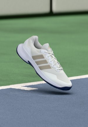 Άσπρο και μπεζ αθλητικό παπούτσι Adidas με ναυτικό μπλε σόλα να αιωρείται πάνω από μια μπλε και πράσινη επιφάνεια γηπέδου τένις.