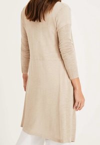 Lång, beige cardigan i ett mjukt, lätt stickat material med lång ärm och en något texturerad yta. Har en medellång design och subtila sömdetaljer.