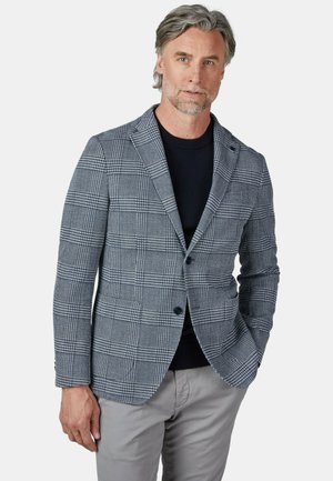 Mann trägt einen grauen karierten Blazer über einem schwarzen Pullover und hellgrauen Hosen, steht mit einer Hand in der Tasche.