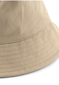 Béžový klobúk s širokou krempou, vyrobený z textúrovanej látky s našitými detailmi. Hladká vnútorná podšívka bez viditeľných log alebo ozdôb.