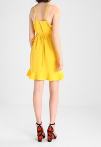 Gelbes Trägerkleid mit gerafftem Saum und einem Bindegürtel in der Taille. Schwarze Absätze mit rotem Blumenmuster auf der Rückseite.