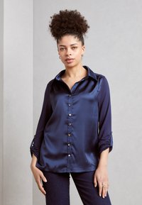 Marineblaues Satinbluse mit spitzem Kragen, langen Ärmeln und Knopfverschluss auf der Vorderseite; verfügt über umgeschlagene Ärmelbündchen und eine glatte Textur.